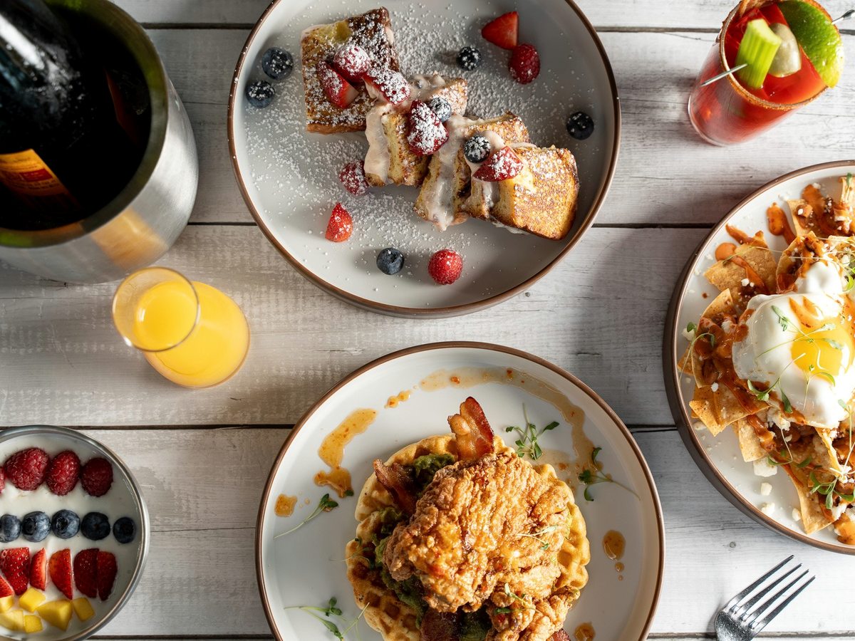 An overhead view of breakfast dishes including fried chicken and waffles, French toast, fresh fruit and orange juice.