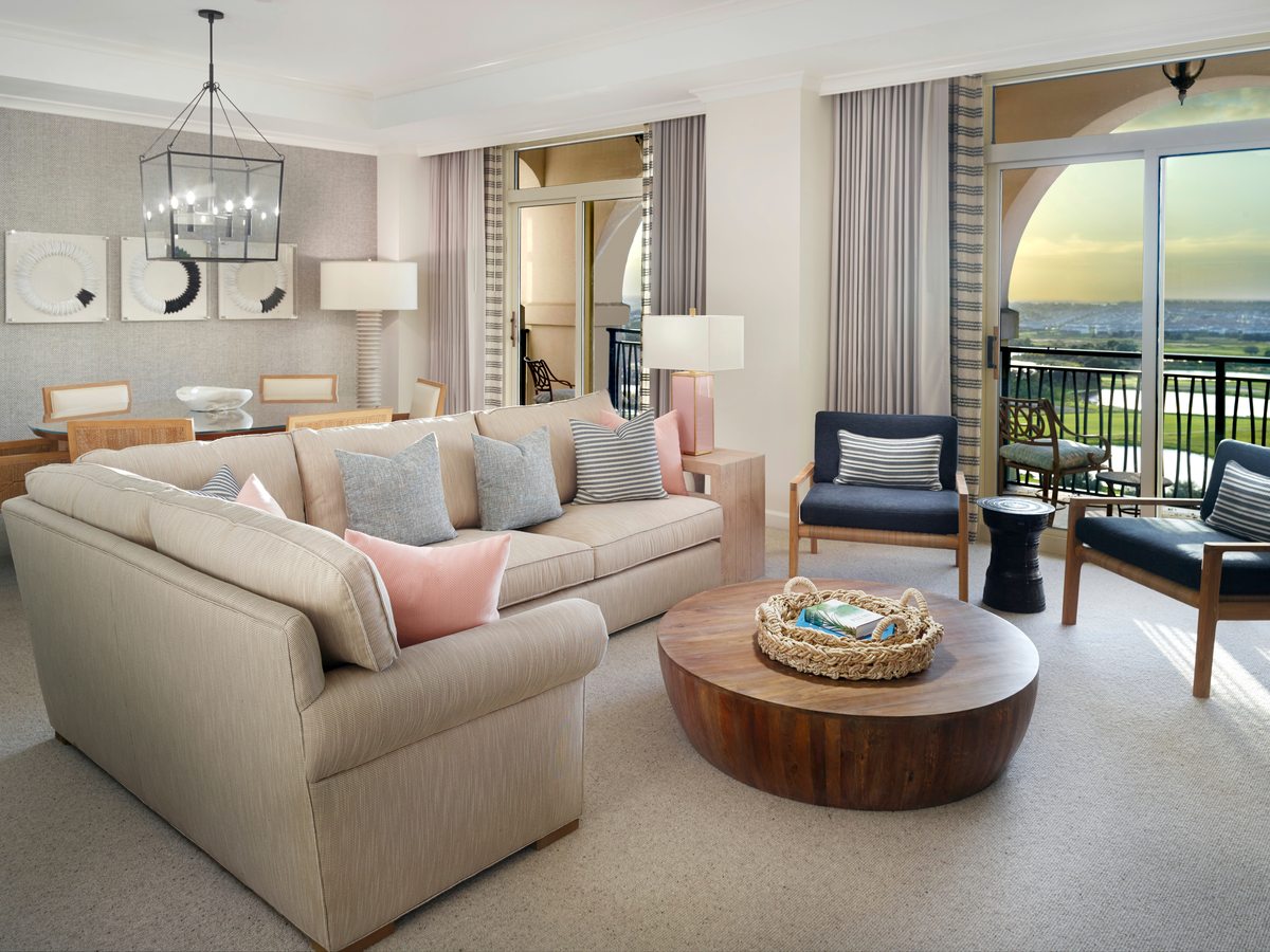An open-plan suite with a cream corner sofa, two armchairs, a round coffee table and a four-seat dining table, with floor-to-ceiling glass onto a balcony and golf view.