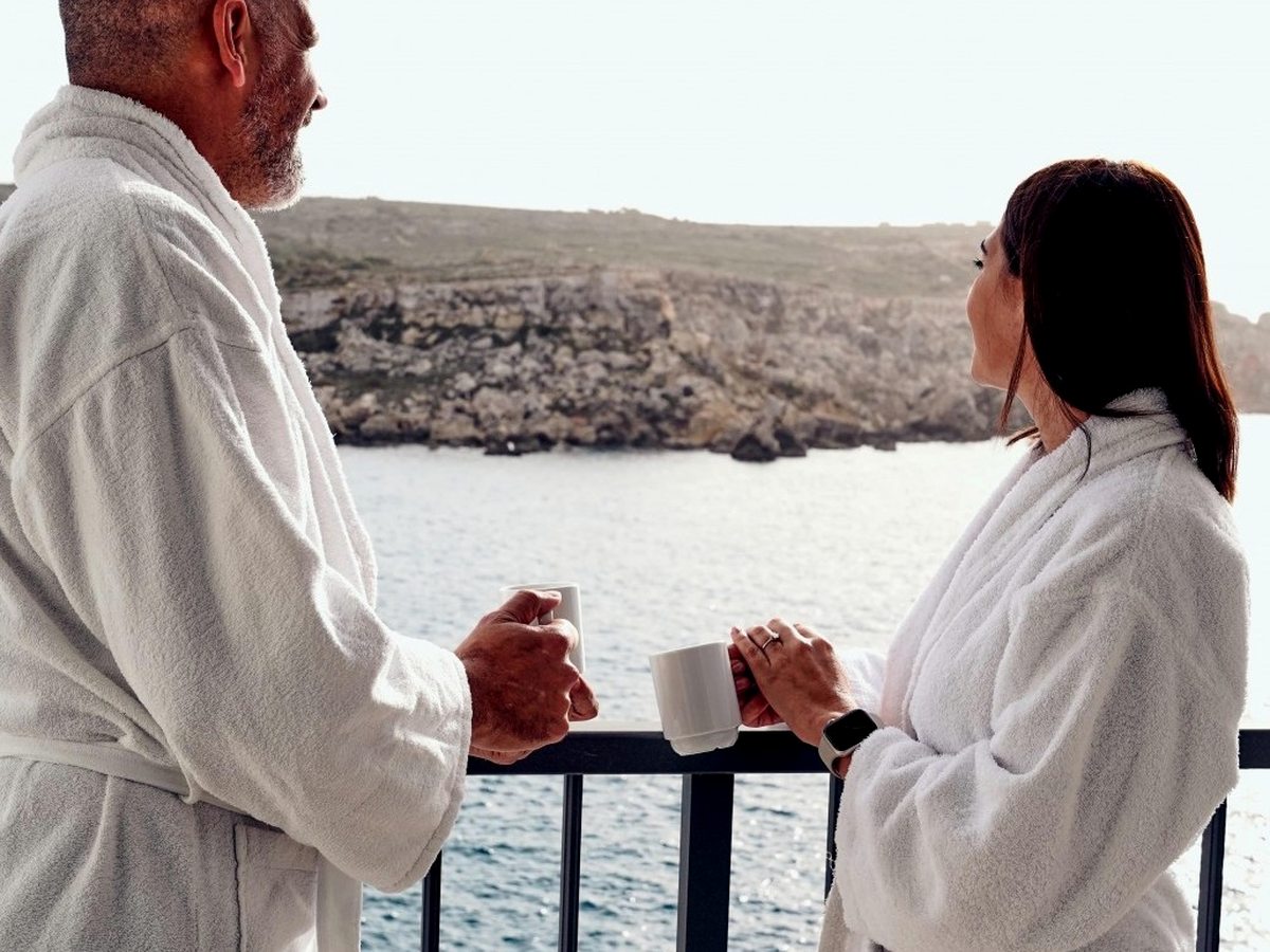 Two guests in white towelling robes standing at a black balcony railing with mugs of coffee, looking across calm blue water to a low rocky headland.