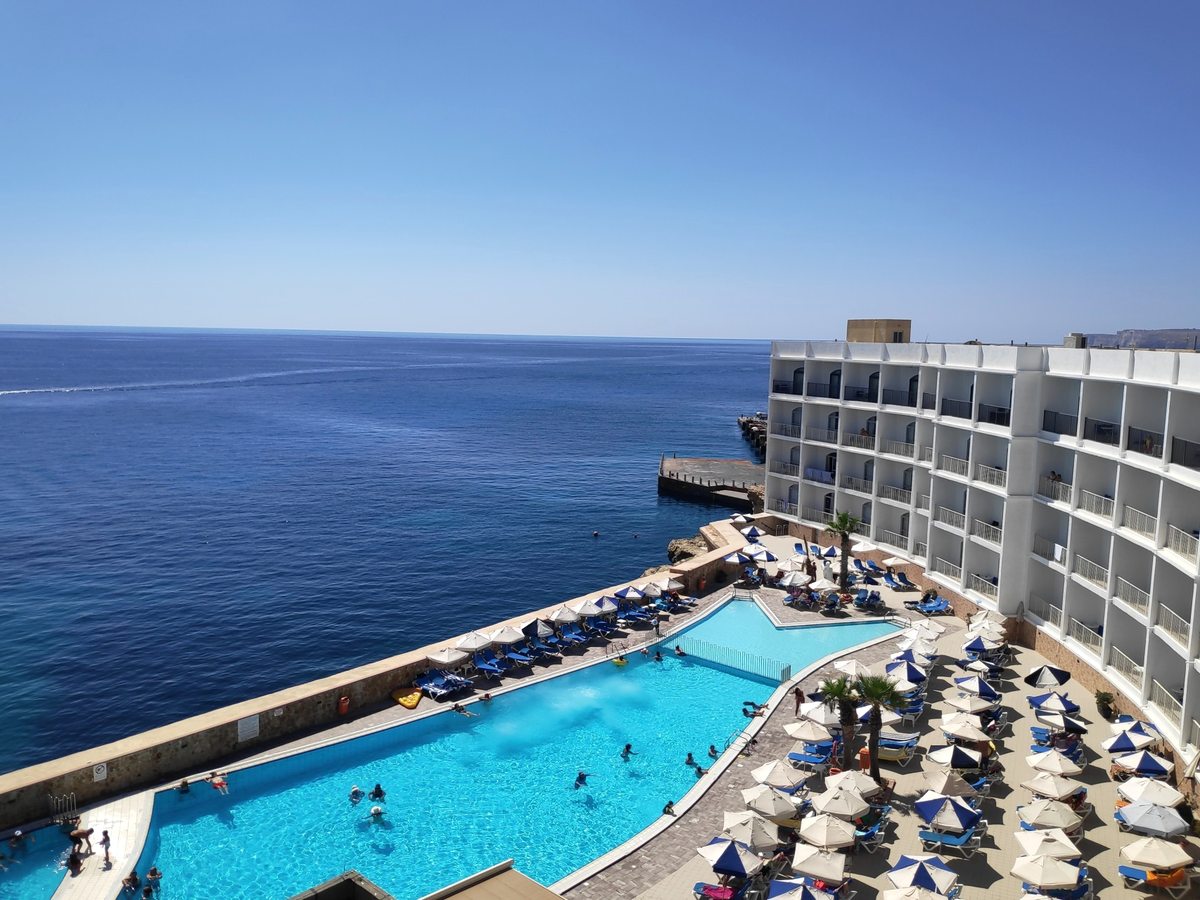 Two connected outdoor pools on a terrace above the sea, edged with rows of blue sun loungers and white parasols, with a wing of the hotel to the right and open water beyond.