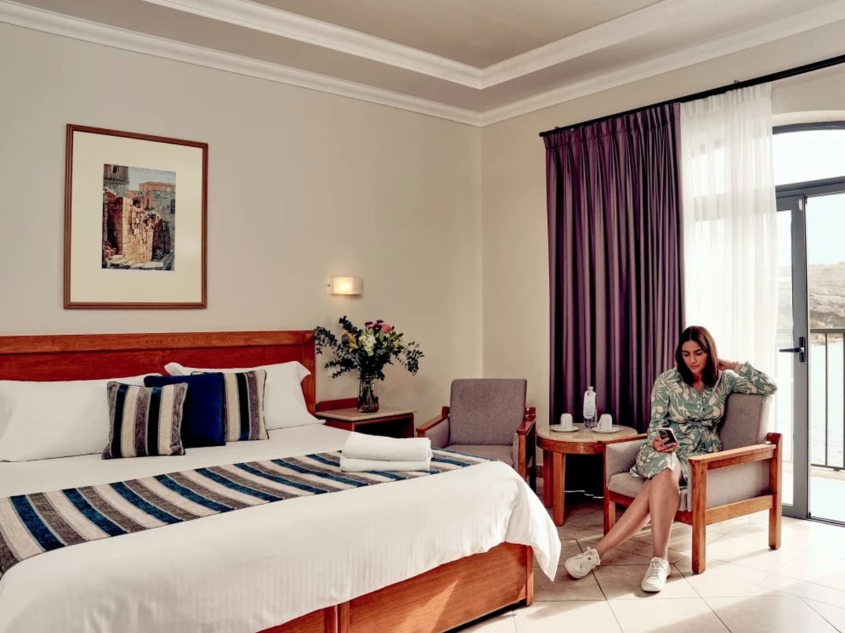 Bright double room with a wooden headboard, blue and grey striped bedding, two grey armchairs and a small round table beside purple curtains and open balcony doors.