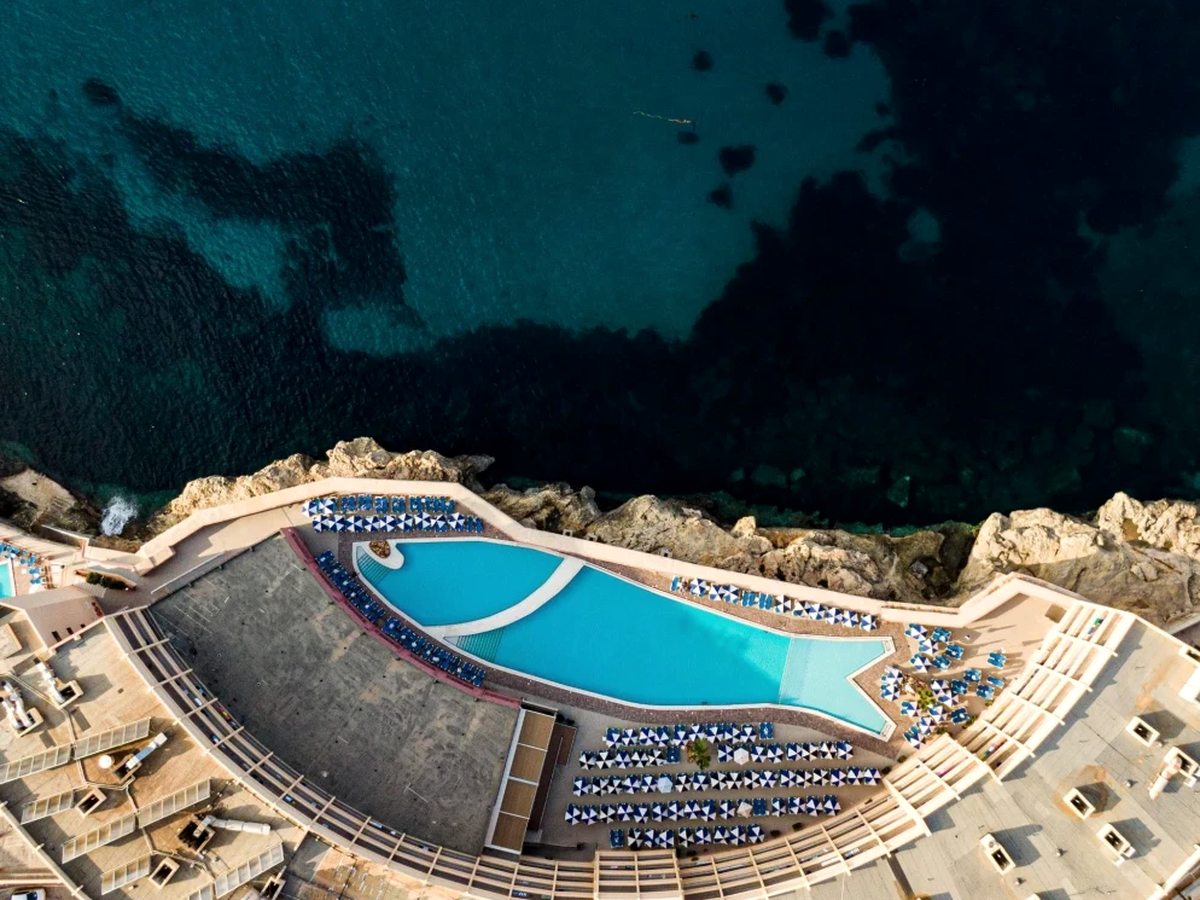 Aerial view looking straight down on the curved outdoor pool and its stone terrace, with lines of blue sun loungers and parasols and the rocky shoreline and dark blue sea alongside.