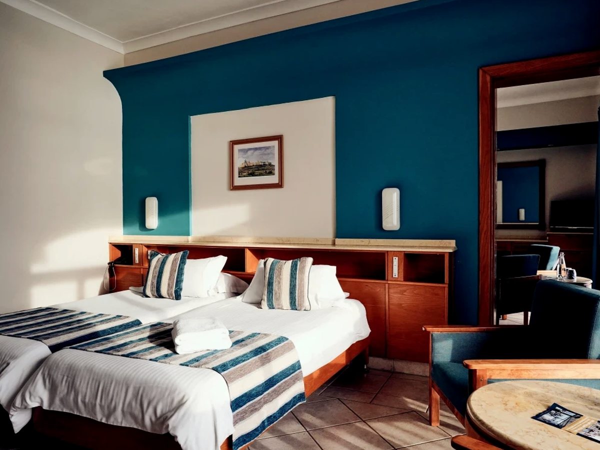Twin room with two single beds pushed together under a wooden headboard shelf, blue and cream striped runners, a teal painted wall, a framed print and armchairs beside a tall mirror.