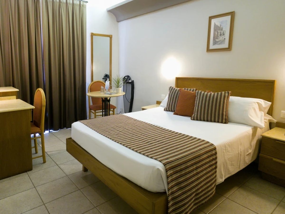 Compact hotel room with a double bed under a pale wooden headboard, a brown striped bed runner, a desk and chair, a tall mirror and floor-length curtains drawn across the balcony doors.