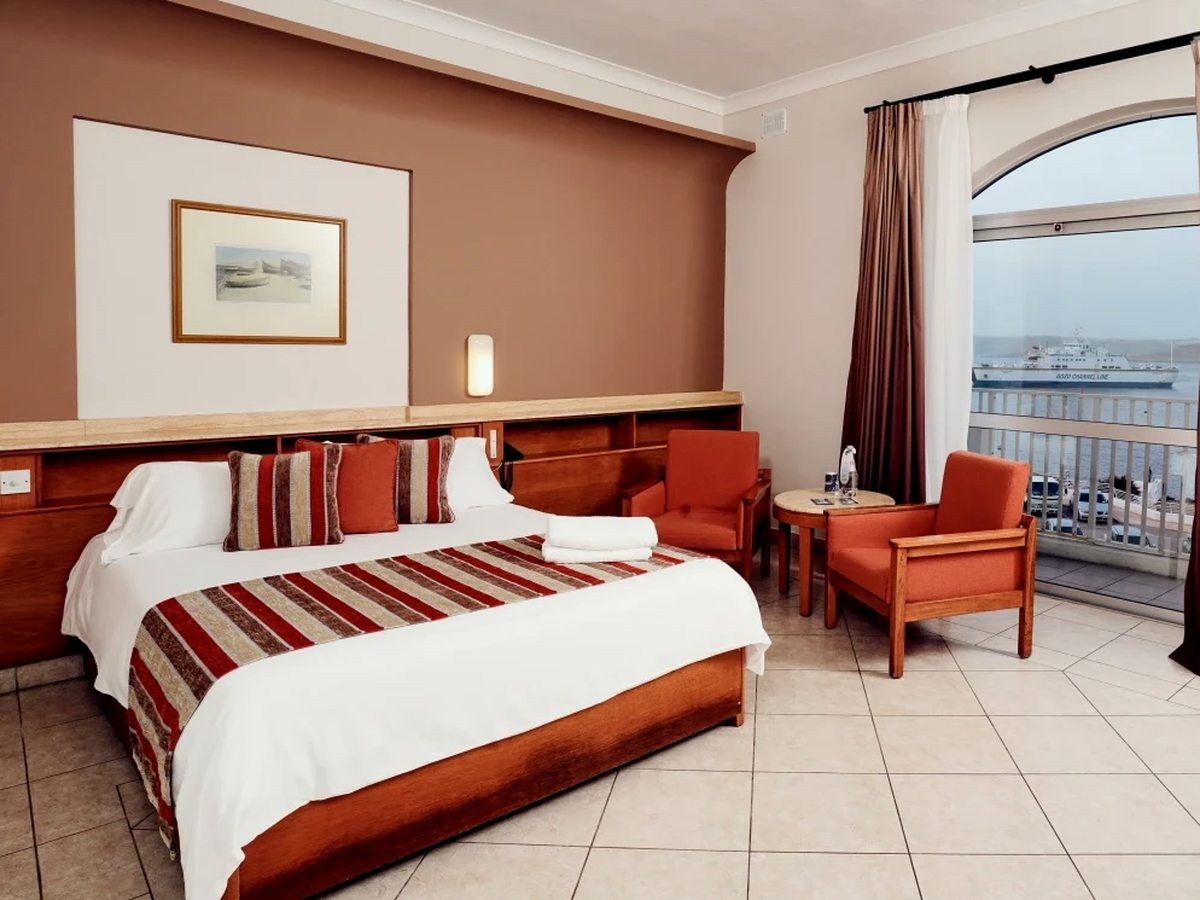 Hotel room with a double bed, a red striped runner and cushions, two orange armchairs and a round table beside an arched balcony door, with a Gozo Channel Line ferry at the terminal outside.