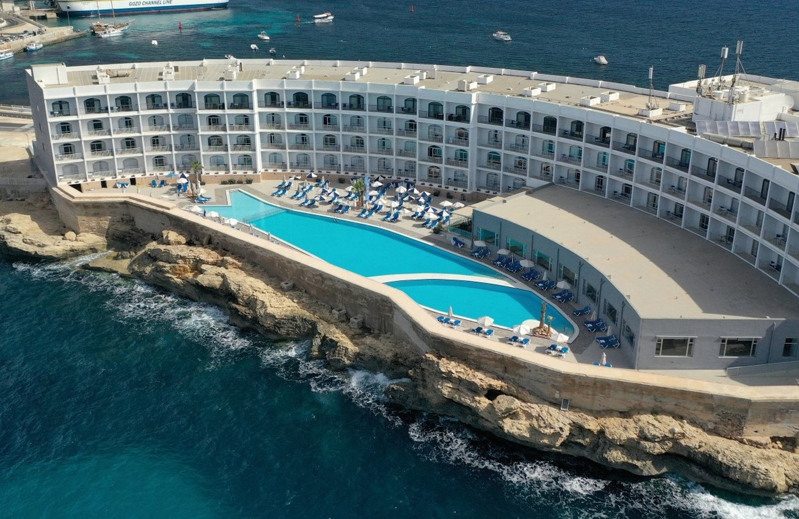 Paradise Bay Resort at Ċirkewwa: a curved white hotel block on the rocky shoreline of Malta's northern tip, with two outdoor pools and rows of blue sun loungers on the terrace below it and a Gozo Channel Line ferry berthed at the terminal beyond.