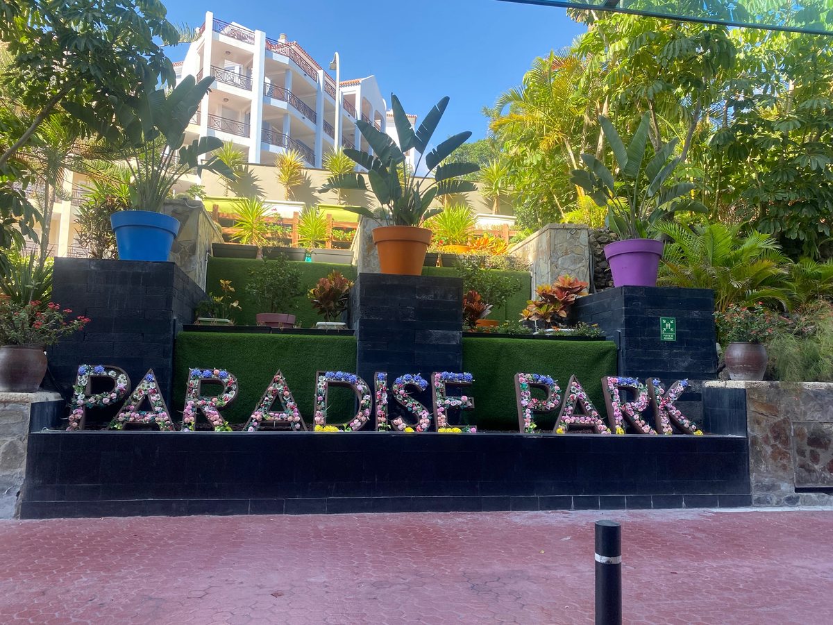 Large PARADISE PARK letters filled with flowers on a black tiled plinth, with coloured plant pots, palms and a white balconied hotel block behind.