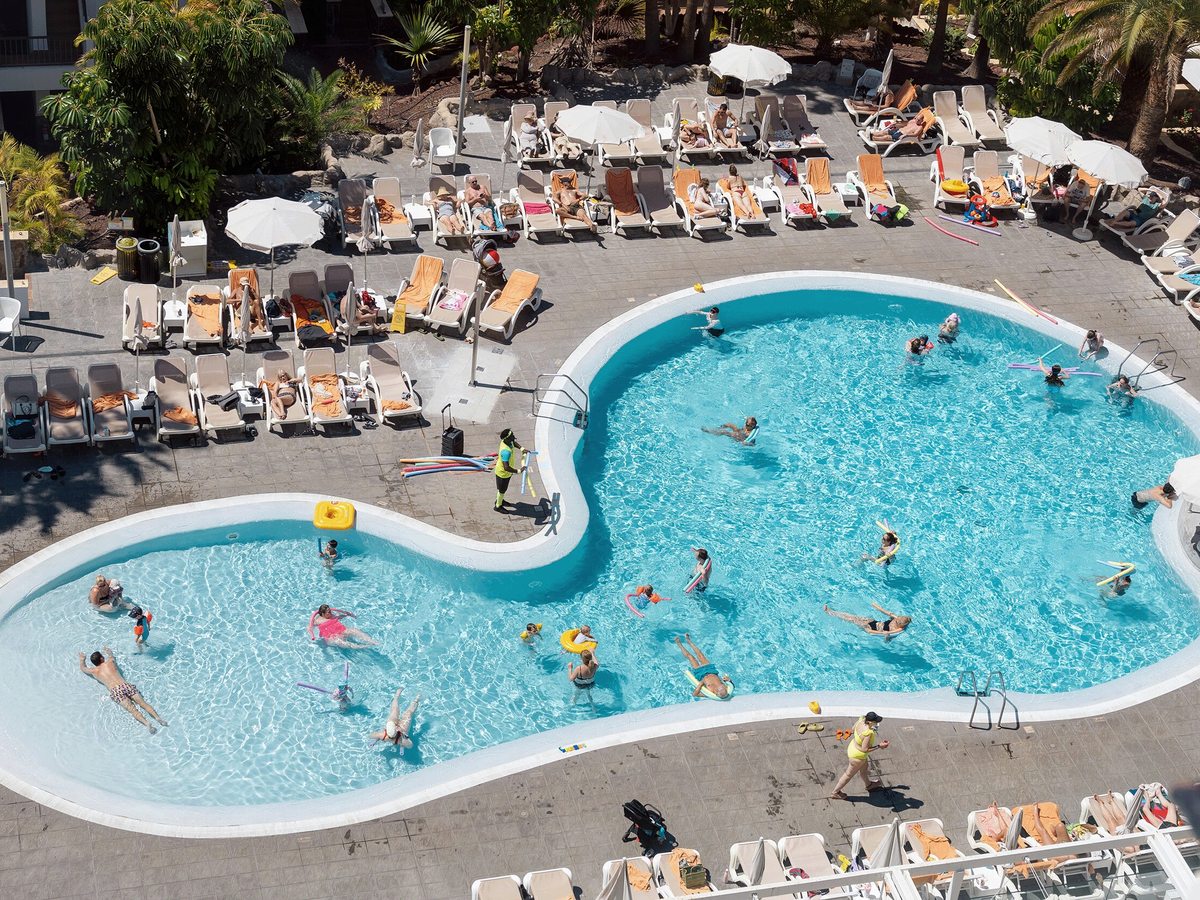 Elevated view of the curved main pool, its shallower lobe on the left with children standing in the water, and rows of sun loungers and parasols on the terrace around it.