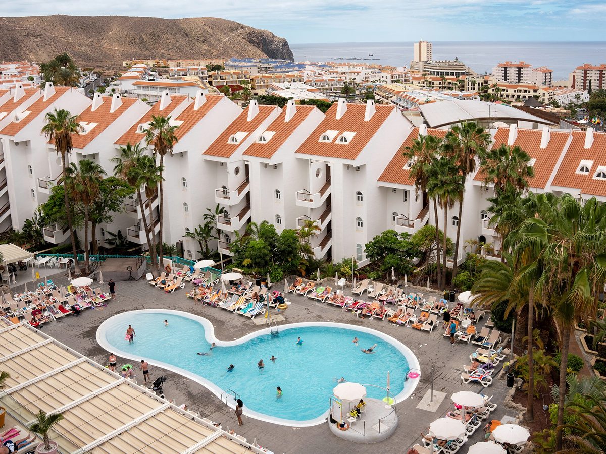 Elevated view over the resort's white blocks with red-tiled roofs and its curved blue pool, with the town of Los Cristianos and the Atlantic beyond.