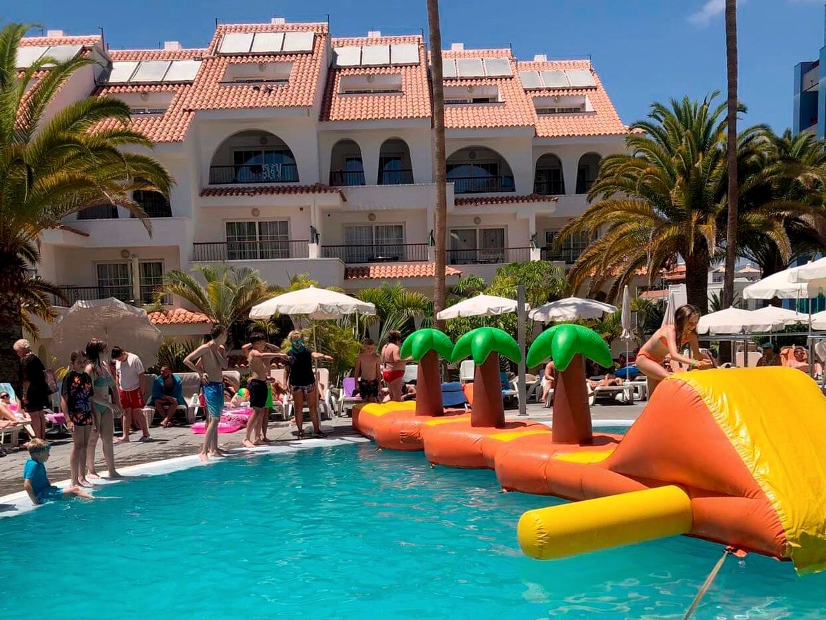 Orange and yellow inflatable obstacle course with green inflatable palm trees floating across the pool, children on the poolside and white balconied blocks behind.