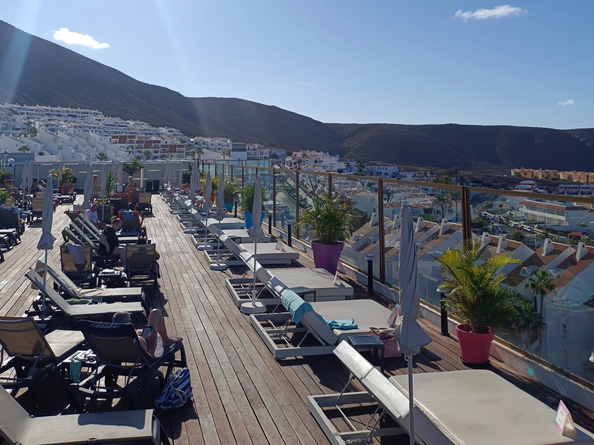 Rows of sun loungers and folded parasols on a timber roof deck behind a glass balustrade, with white houses and a bare volcanic ridge beyond.