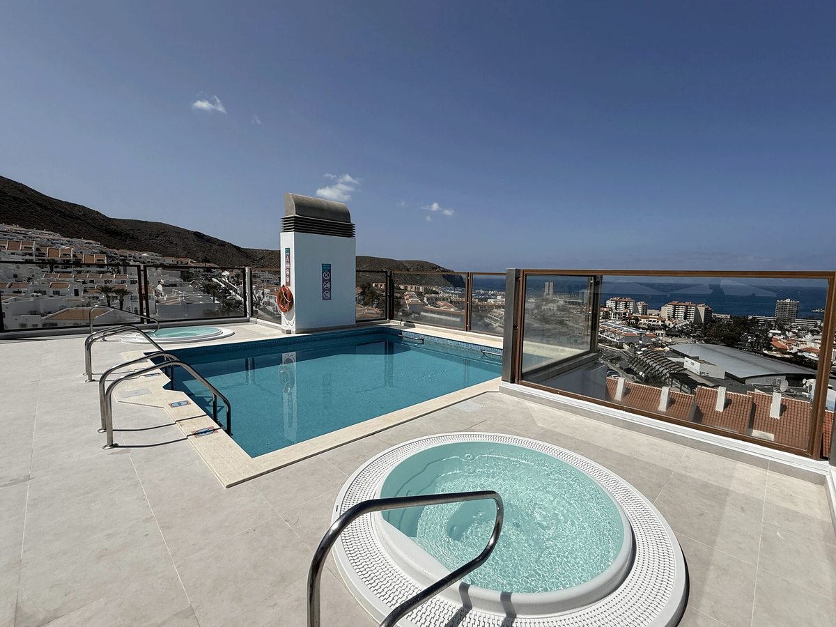Small rectangular rooftop pool with a circular whirlpool in front of it on a pale tiled terrace, a glass balustrade and the town and hills beyond.
