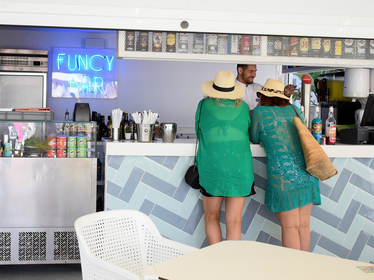 Two guests in sun hats ordering at a pale tiled bar counter under a blue neon FunCy Bar sign, with a member of staff serving and bottles and juice machines behind.