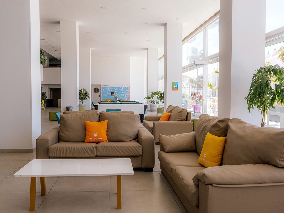 Bright open lounge with tan leather sofas, orange and yellow cushions and a white coffee table, with a pool table and a noticeboard at the far end.