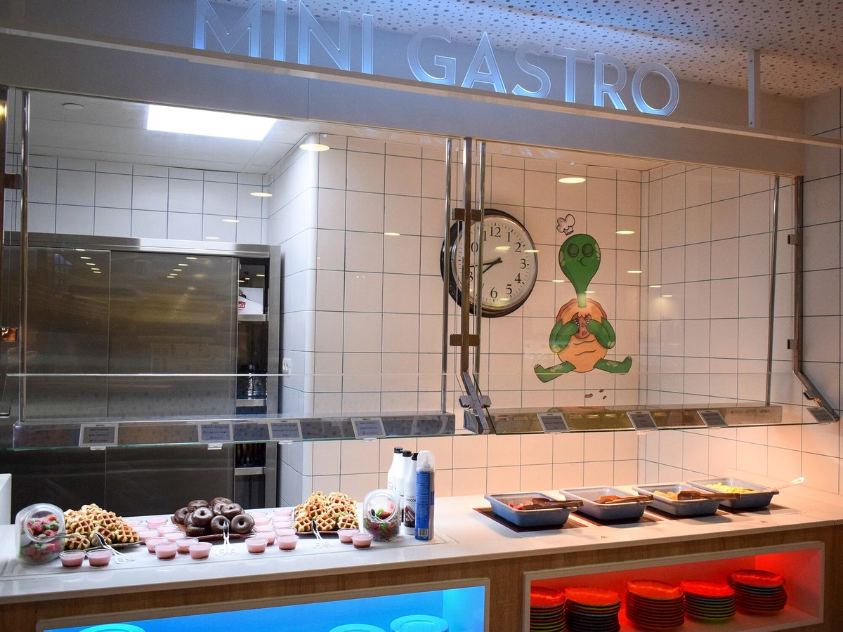 Low white food counter under a MINI GASTRO sign, laid with waffles, chocolate doughnuts and small pink desserts, with hot trays and stacks of red plates alongside.