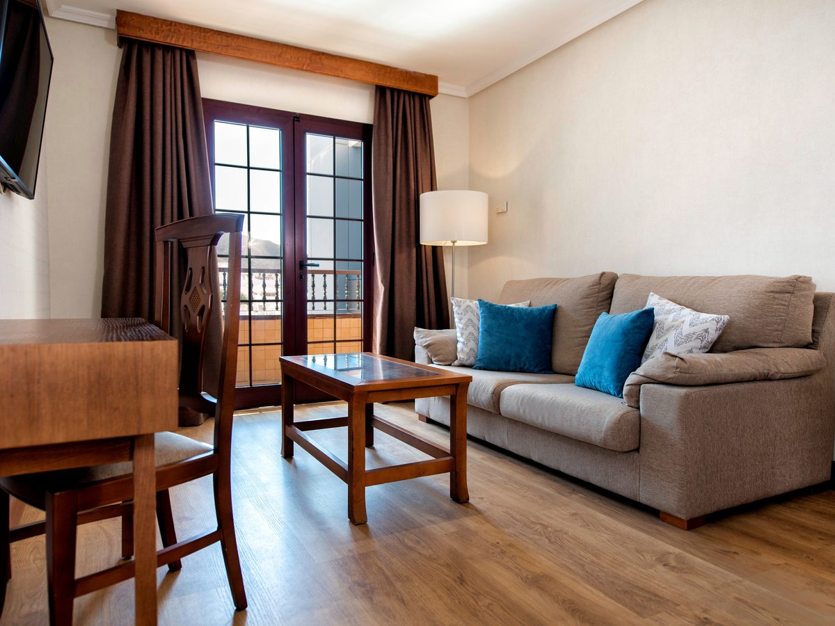 Living room with a grey sofa and blue cushions, a wooden coffee table, a dining table and chairs and a wall-mounted television, with French doors onto a balcony.