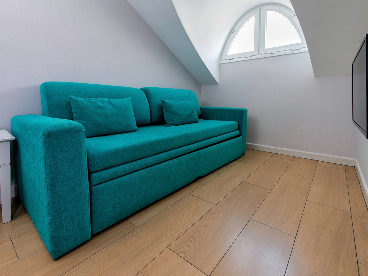 Turquoise sofa bed against a wall beneath a sloping ceiling and a small arched window, on a wood-effect floor.