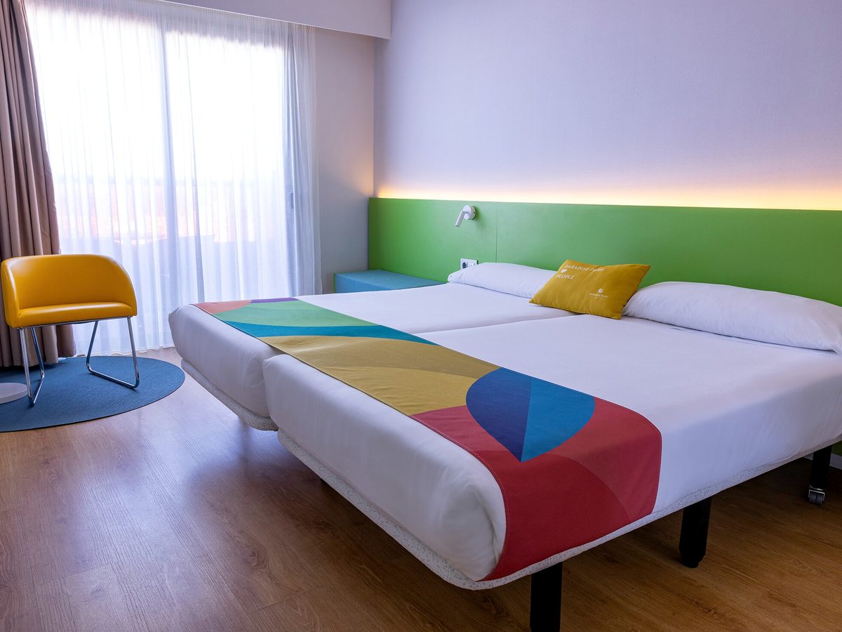 Twin beds pushed together under a green headboard with a multicoloured bed runner and a yellow cushion, a yellow armchair on a blue round rug beside curtained balcony doors.