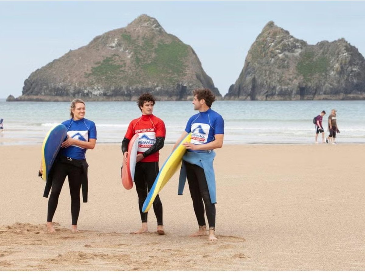 Three people in wetsuits and blue rash vests carrying foam surfboards on wide golden sand, with two tall green-topped rock stacks rising out of the sea behind them.