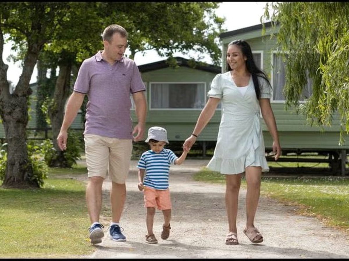 A man, a woman and a young boy in a sun hat walking hand in hand along a gravel path past pale green static caravans, with lawns, a willow and mature trees around them.