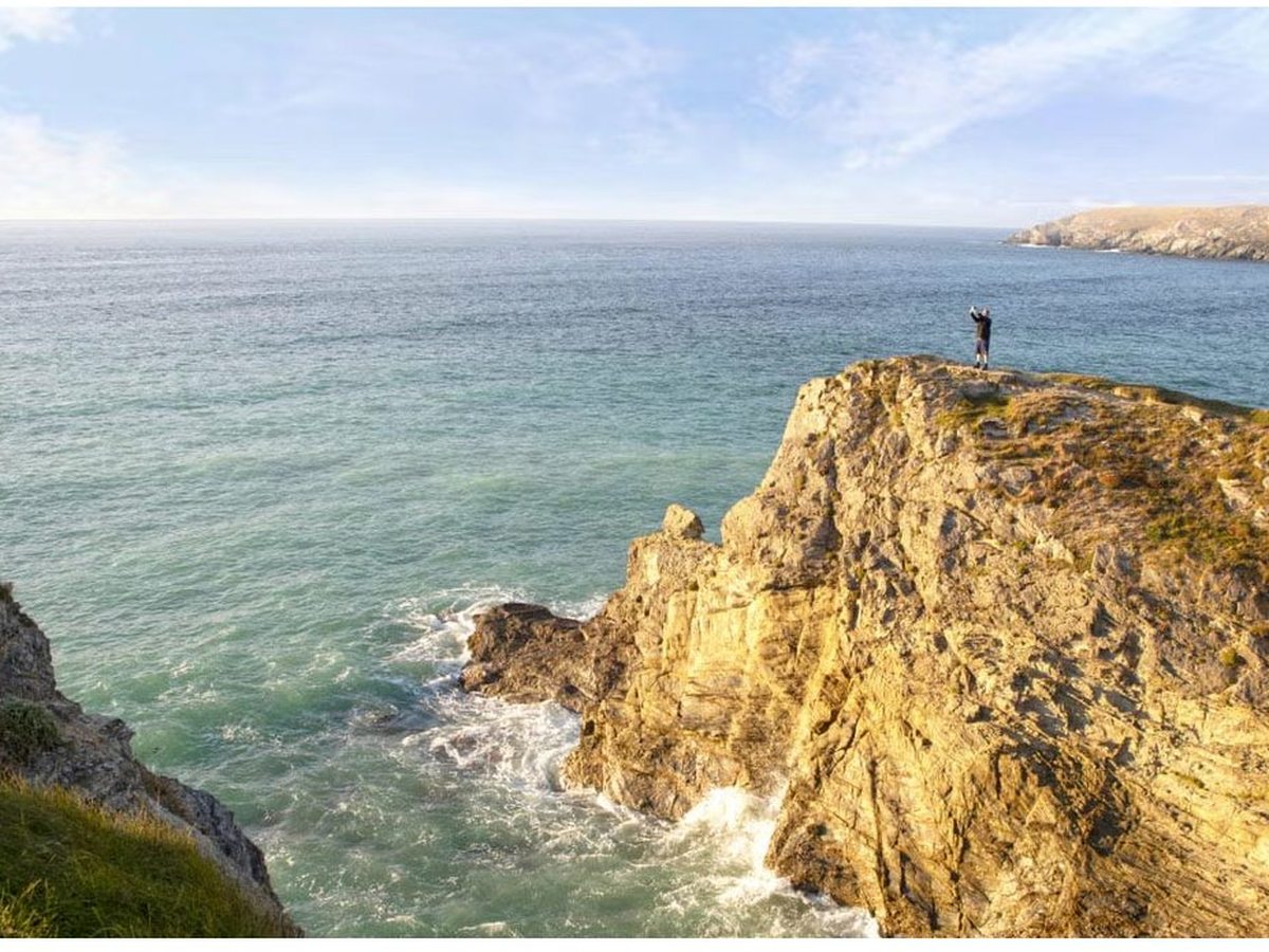A walker standing on a golden rocky headland above the sea, with waves breaking on the rocks below and further cliffs along the coast beyond.