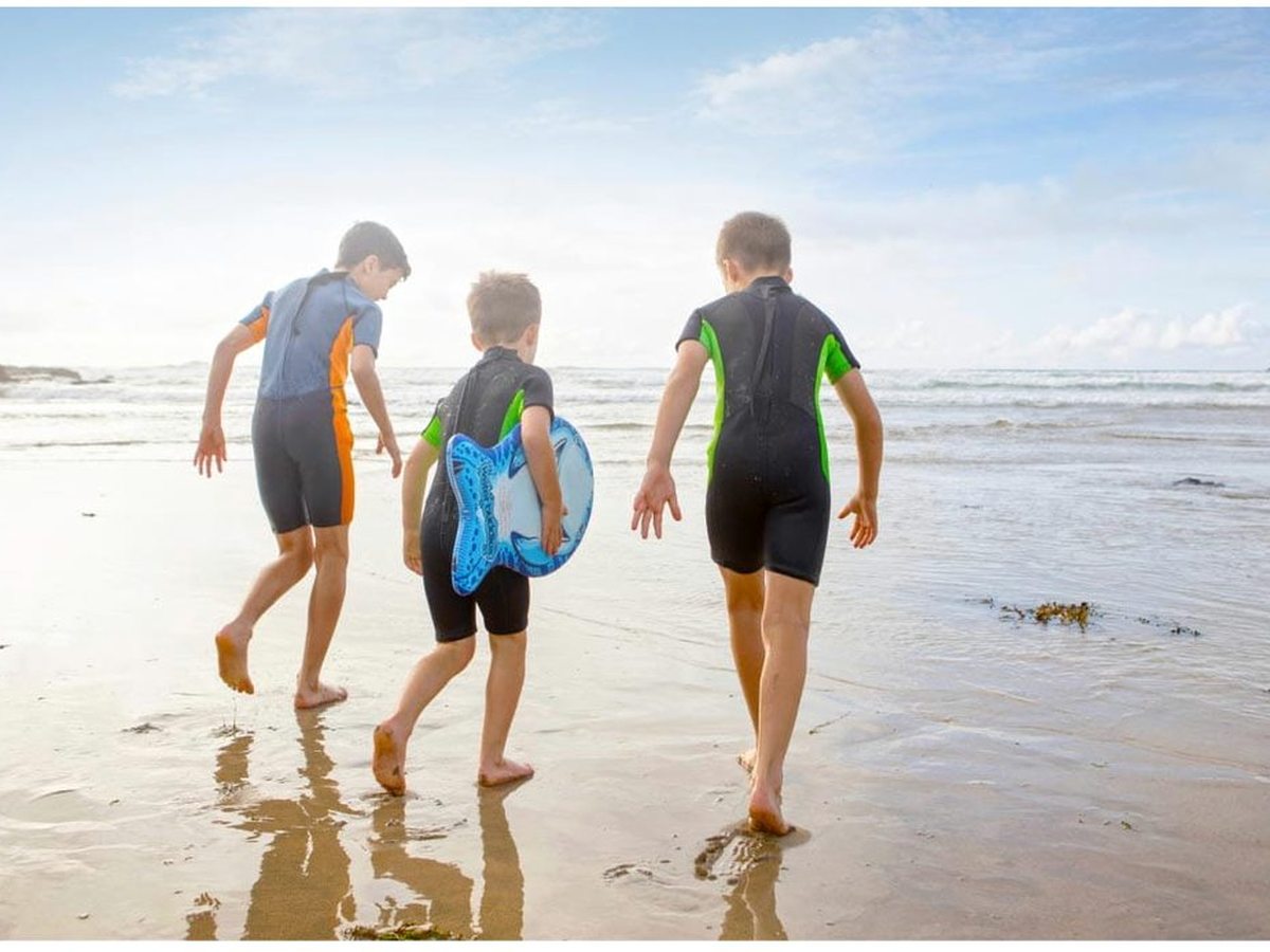 Three boys in short wetsuits walking across wet, reflective sand towards the breaking waves, the middle one carrying a blue bodyboard.