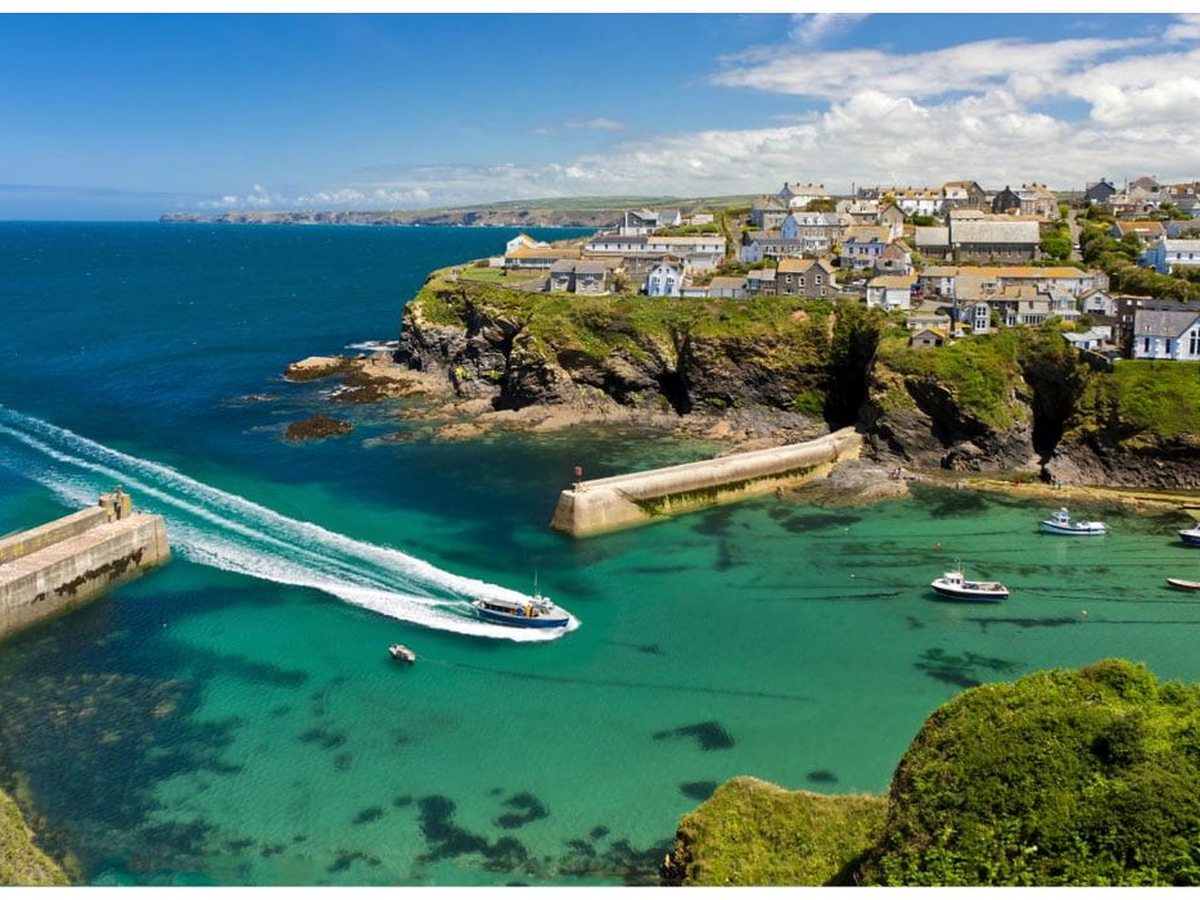 A small stone-walled harbour with two breakwaters and moored fishing boats in clear turquoise water, below a village of slate-roofed cottages on a green headland.