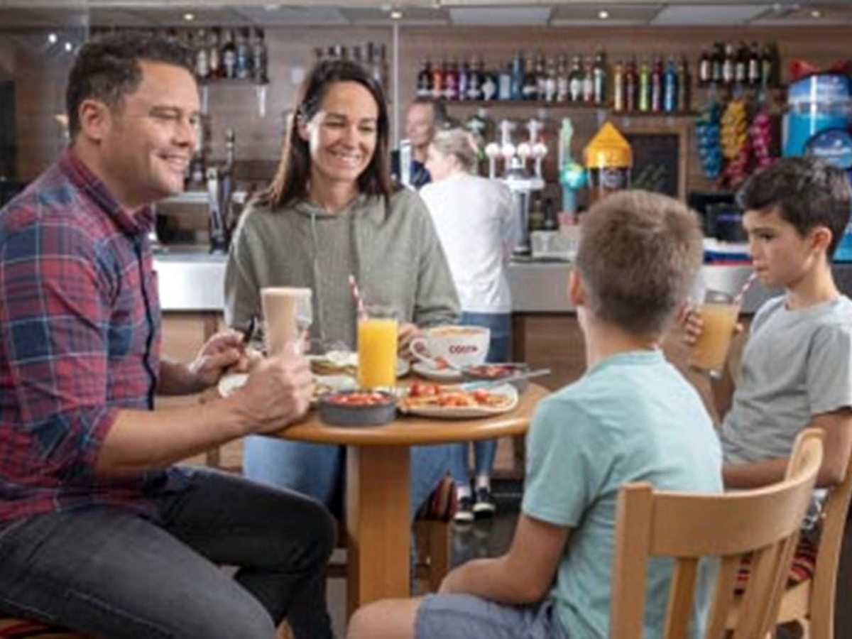 A man, a woman and two boys sitting at a round wooden table with coffee, orange juice and plates of food, in front of a bar counter stocked with bottles and beer taps.