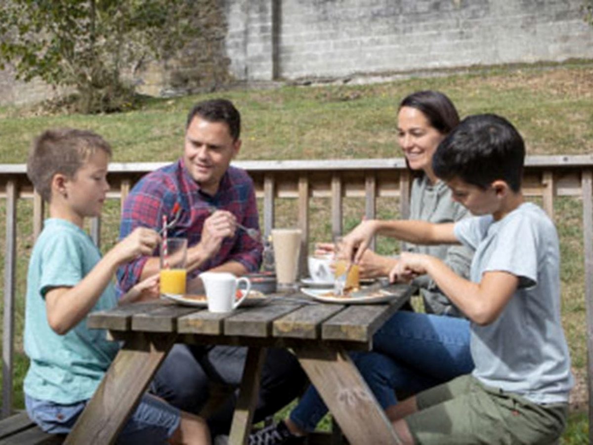 Two adults and two boys eating a cooked breakfast at a wooden picnic table on decking, with a timber rail, grass and a stone wall behind them.
