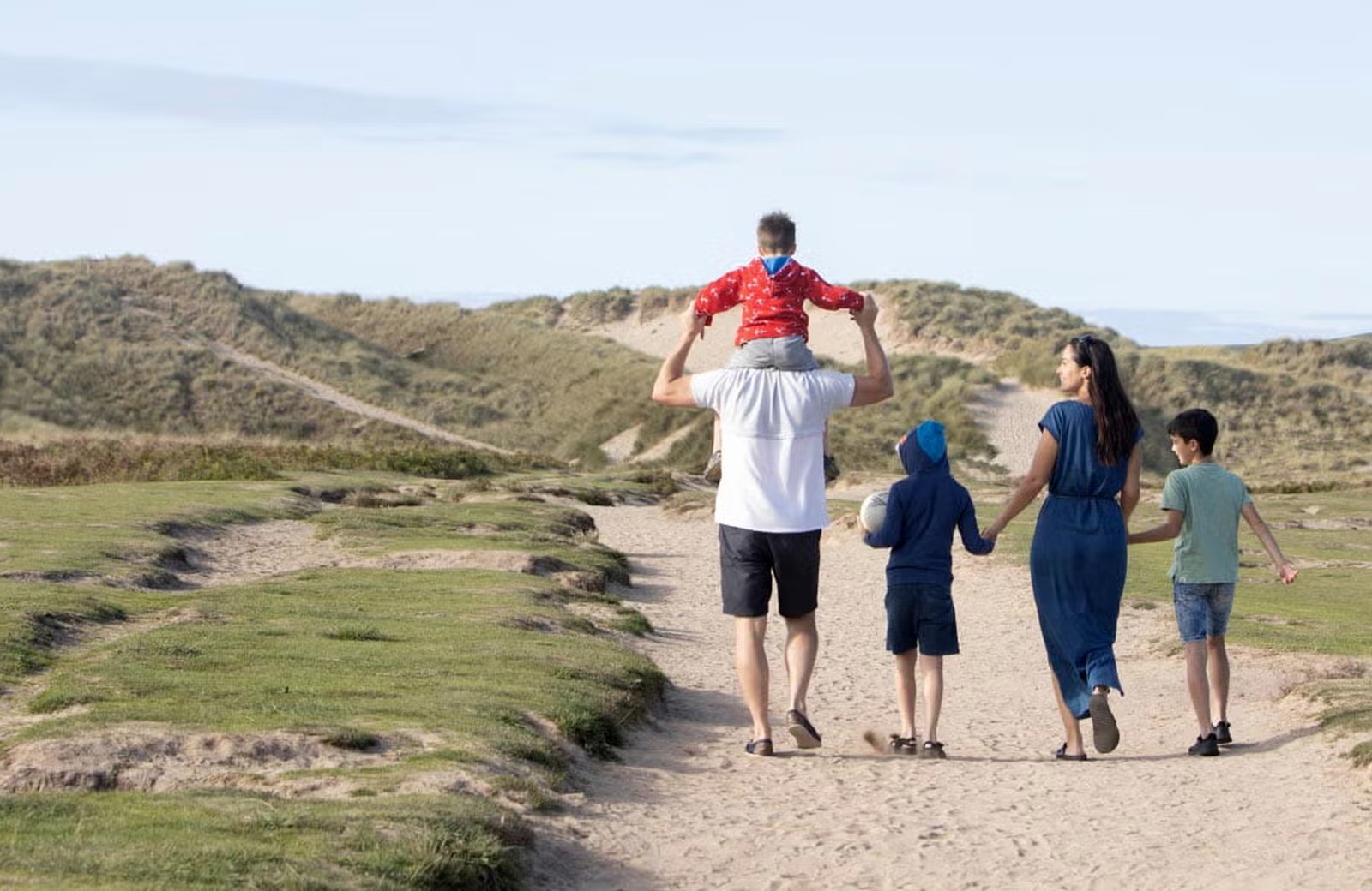 A family walking a sandy path through grassy dunes towards the sea, the youngest child riding on his father's shoulders.