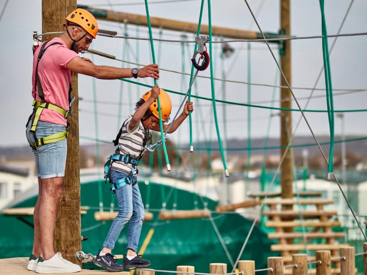 A harnessed and helmeted child stepping along a rope bridge on an outdoor high ropes course, steadied by a helmeted adult on the platform, with caravans behind.