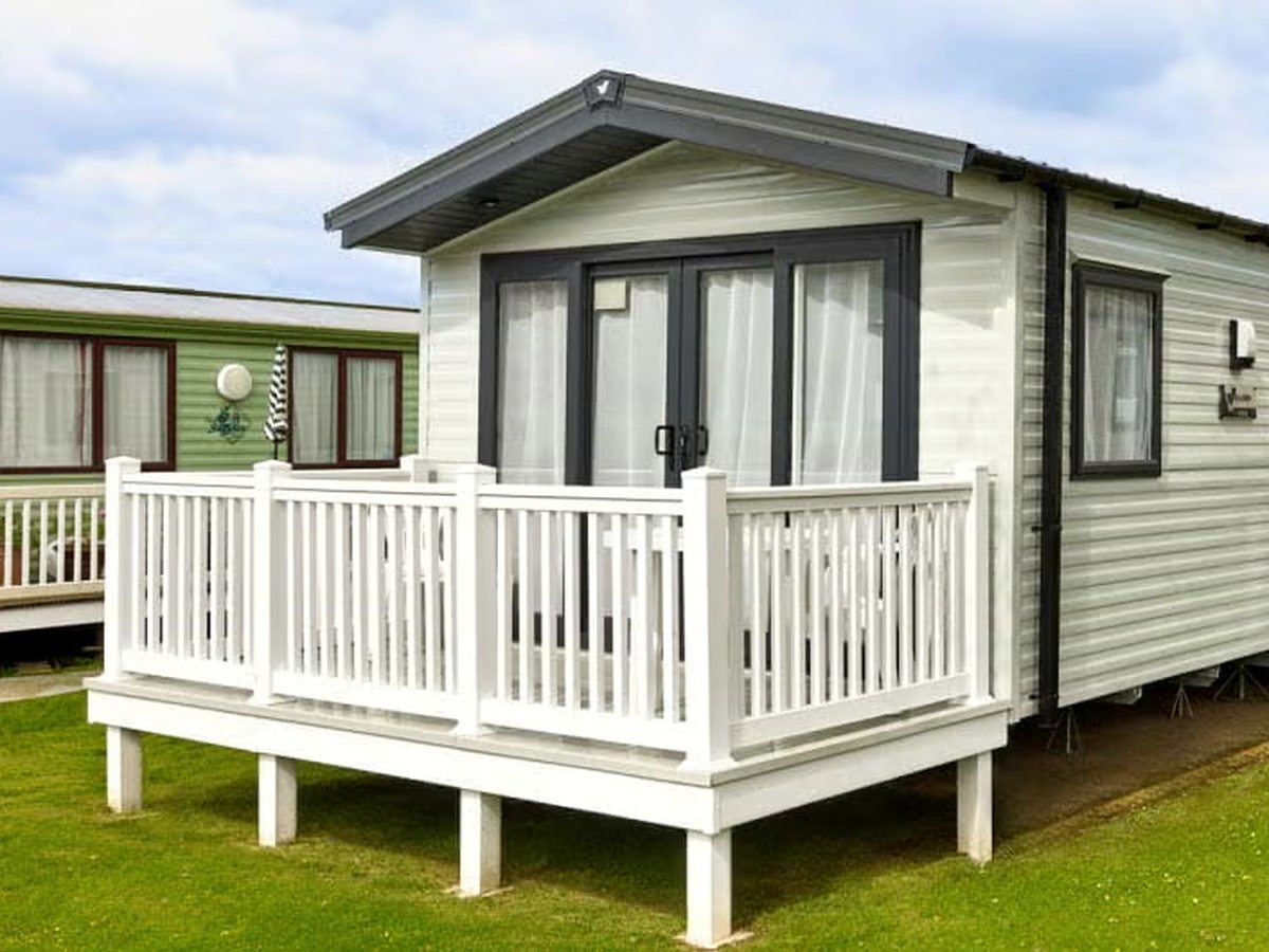 A white static caravan with patio doors and curtained windows, fronted by raised decking with white railings, on a grass pitch with neighbouring caravans behind.