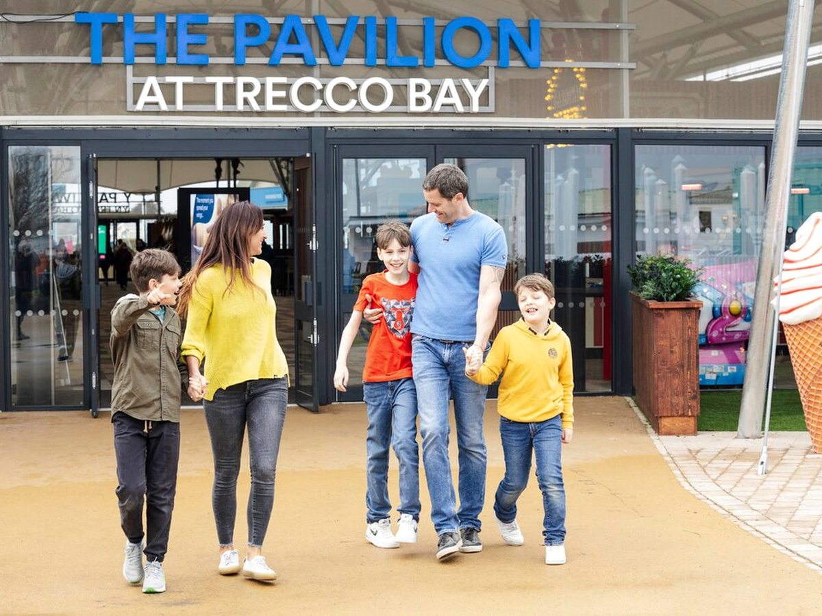 Two adults and three children walking out through the glass doors of the Pavilion, the venue name lettered above them and a giant ice-cream model beside the paved approach.