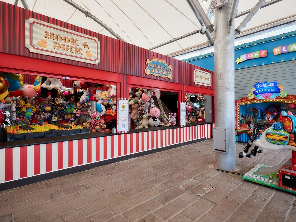 A red and white striped carnival stall with Hook a Duck and basketball games hung with plush prizes, beside the lit entrance to the soft play.