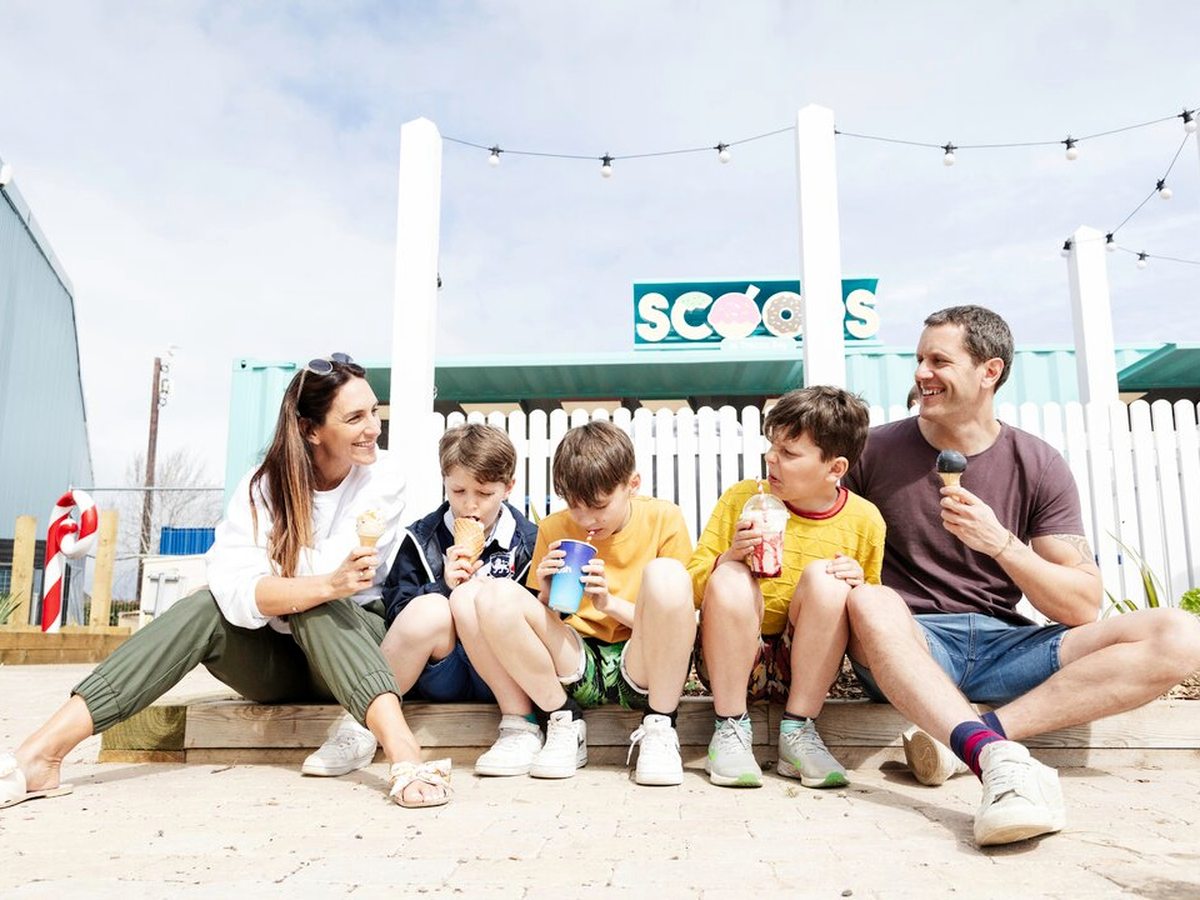Two adults and three children sitting on timber steps with ice creams and milkshakes, in front of the turquoise Scoops kiosk and its festoon lights.
