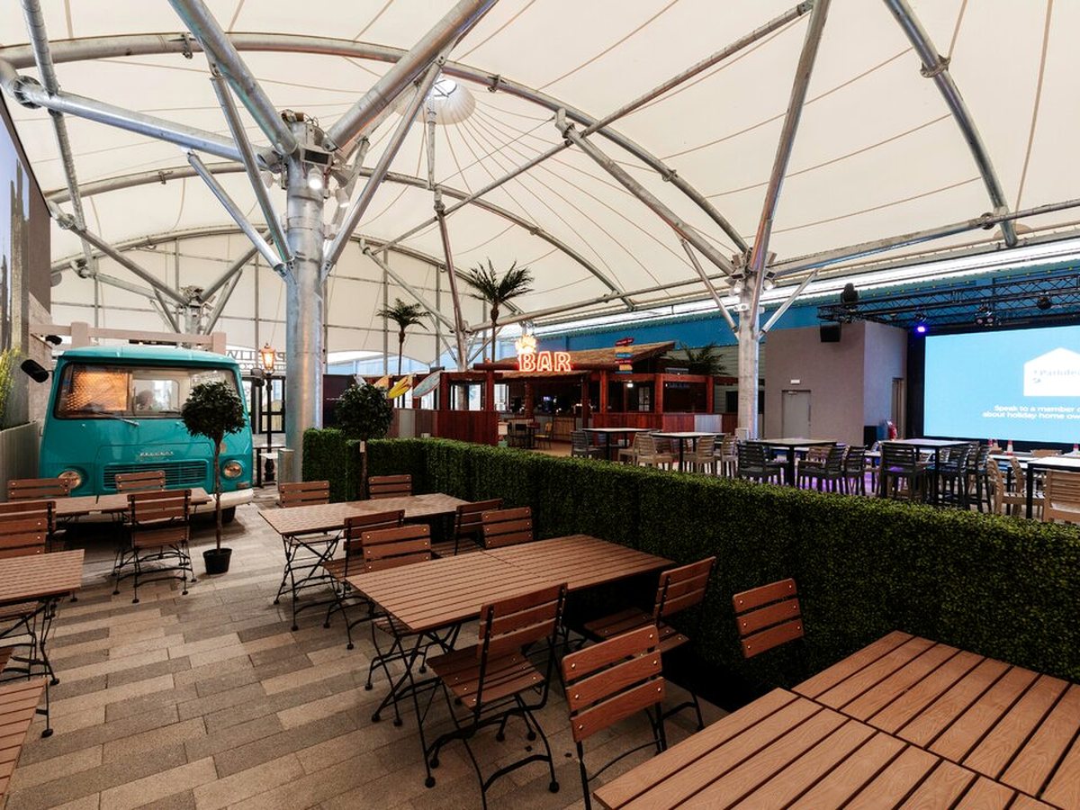 A covered seating area under a white tensile canopy, with wooden tables and benches, a turquoise food van, a bar counter and a large screen at the far end.