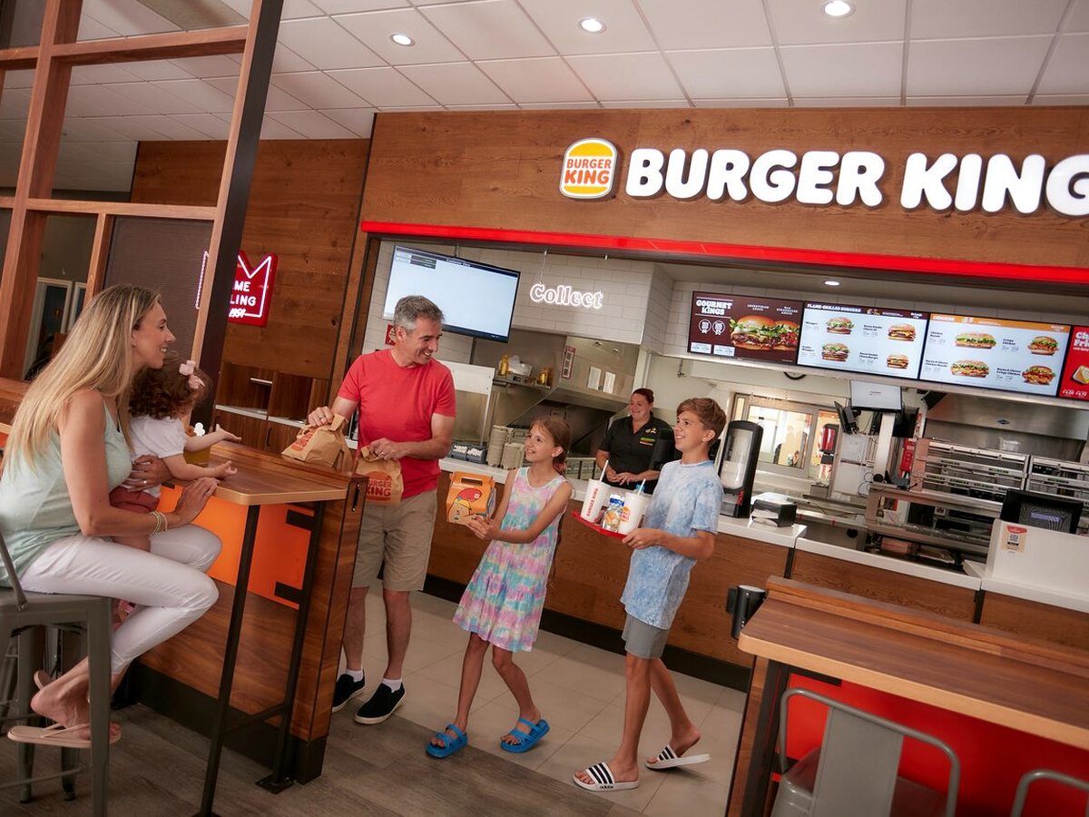 A family collecting takeaway bags at the counter of a Burger King outlet, with the illuminated menu boards and open kitchen behind them.
