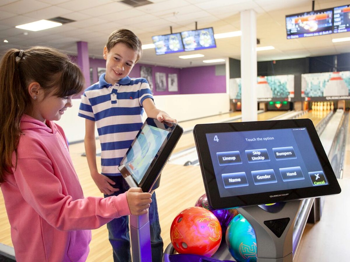 Two children at a touchscreen scoring kiosk beside a rack of bowling balls, with the lanes and overhead screens behind them.