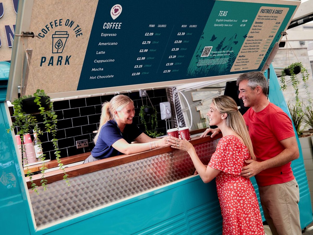 A barista handing a takeaway coffee across a turquoise counter to two customers, with the coffee and tea menu boards above.