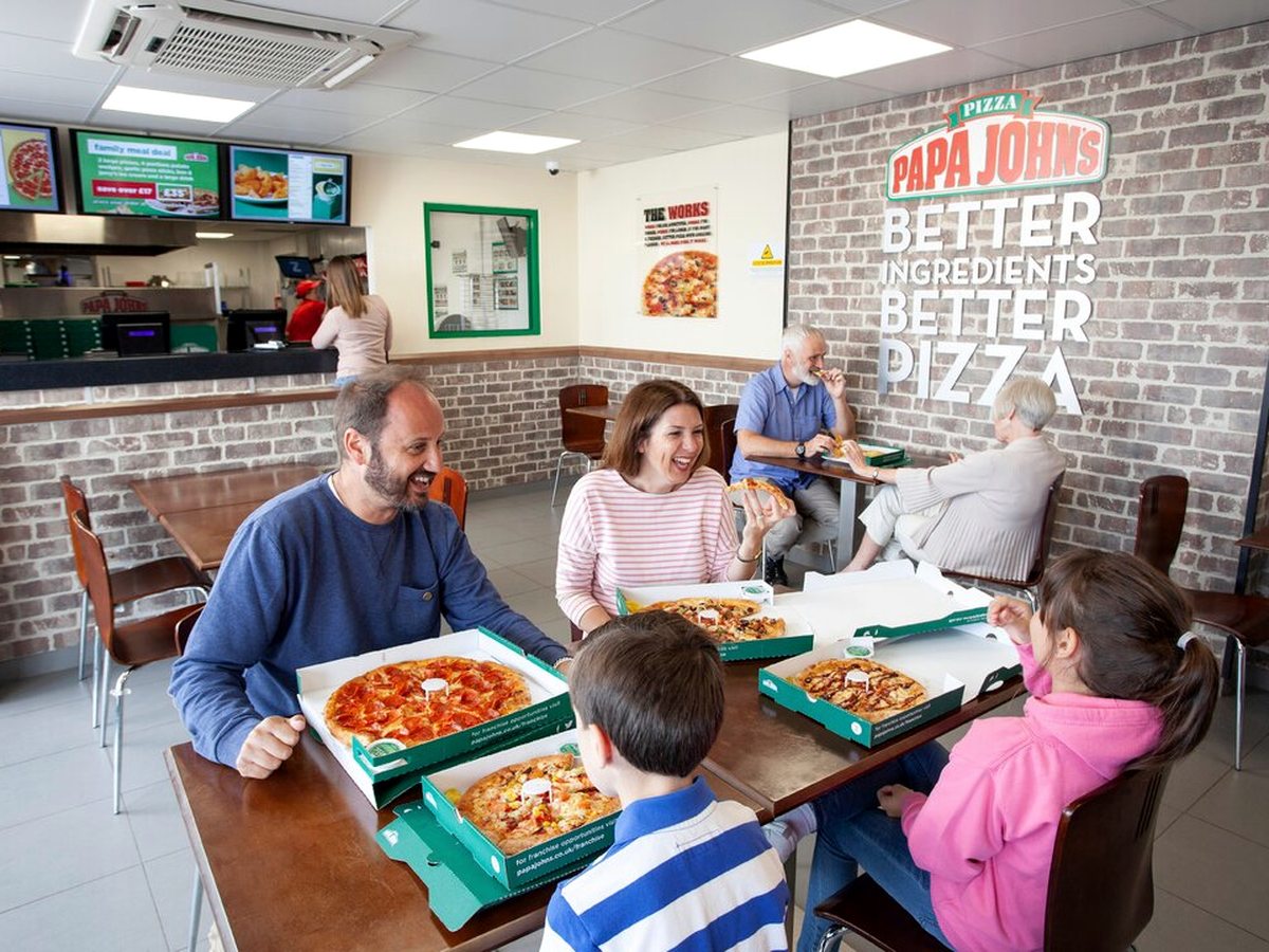 Two families eating pizza at wooden tables in a brick-walled Papa John's, with the service counter and menu screens behind them.