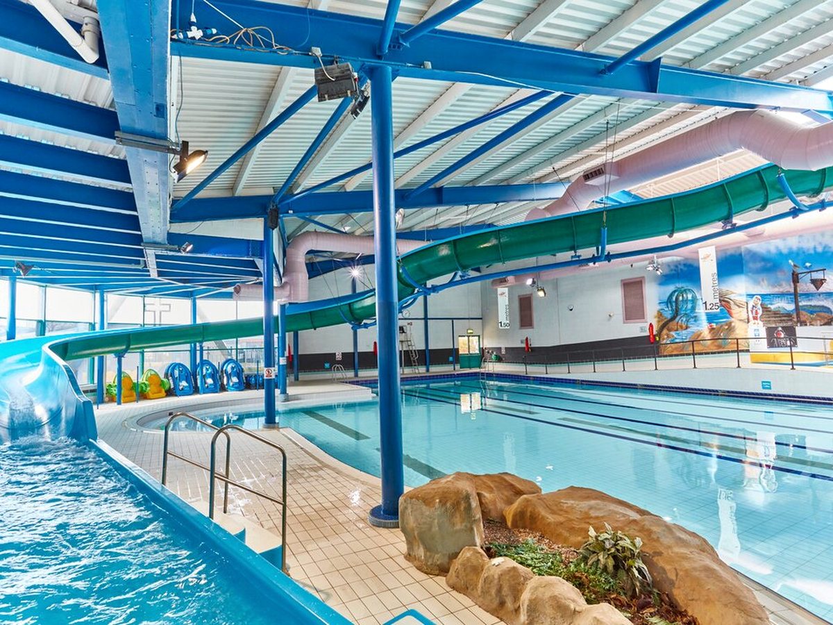 An indoor pool hall under a steel roof with blue trusses, a green enclosed flume curving down to a blue open chute beside rock-effect landscaping and stainless steel handrails.