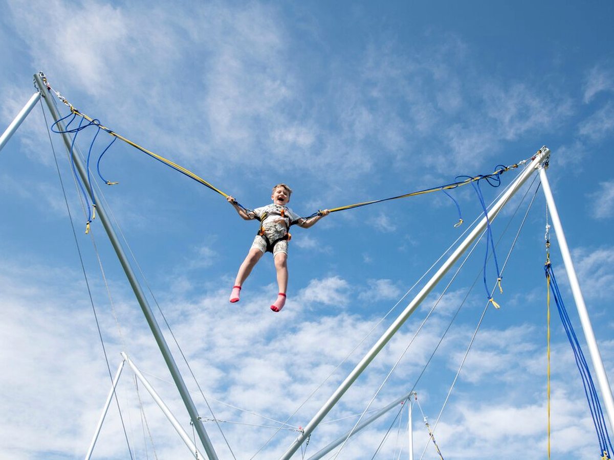A harnessed child bouncing high between the elastic cords of a bungee trampoline against a blue sky.