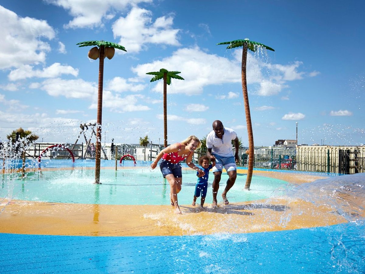 Two adults and a toddler running through shallow water on a splash pad, with palm-tree water features spraying overhead under a blue sky.