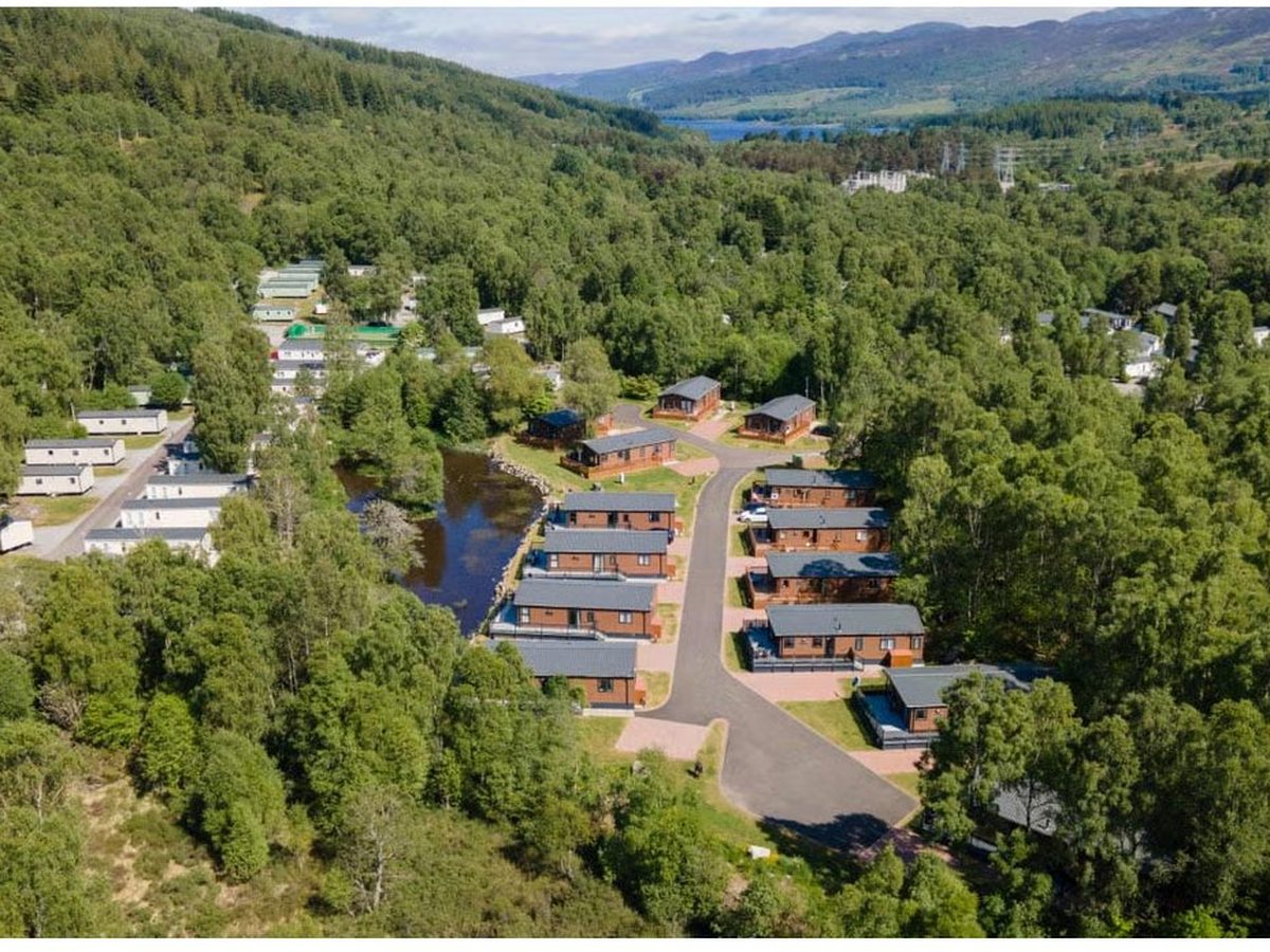 Aerial view of the holiday park, with lodges and static caravans along winding internal roads through dense forest in a Highland valley, a loch in the distance.