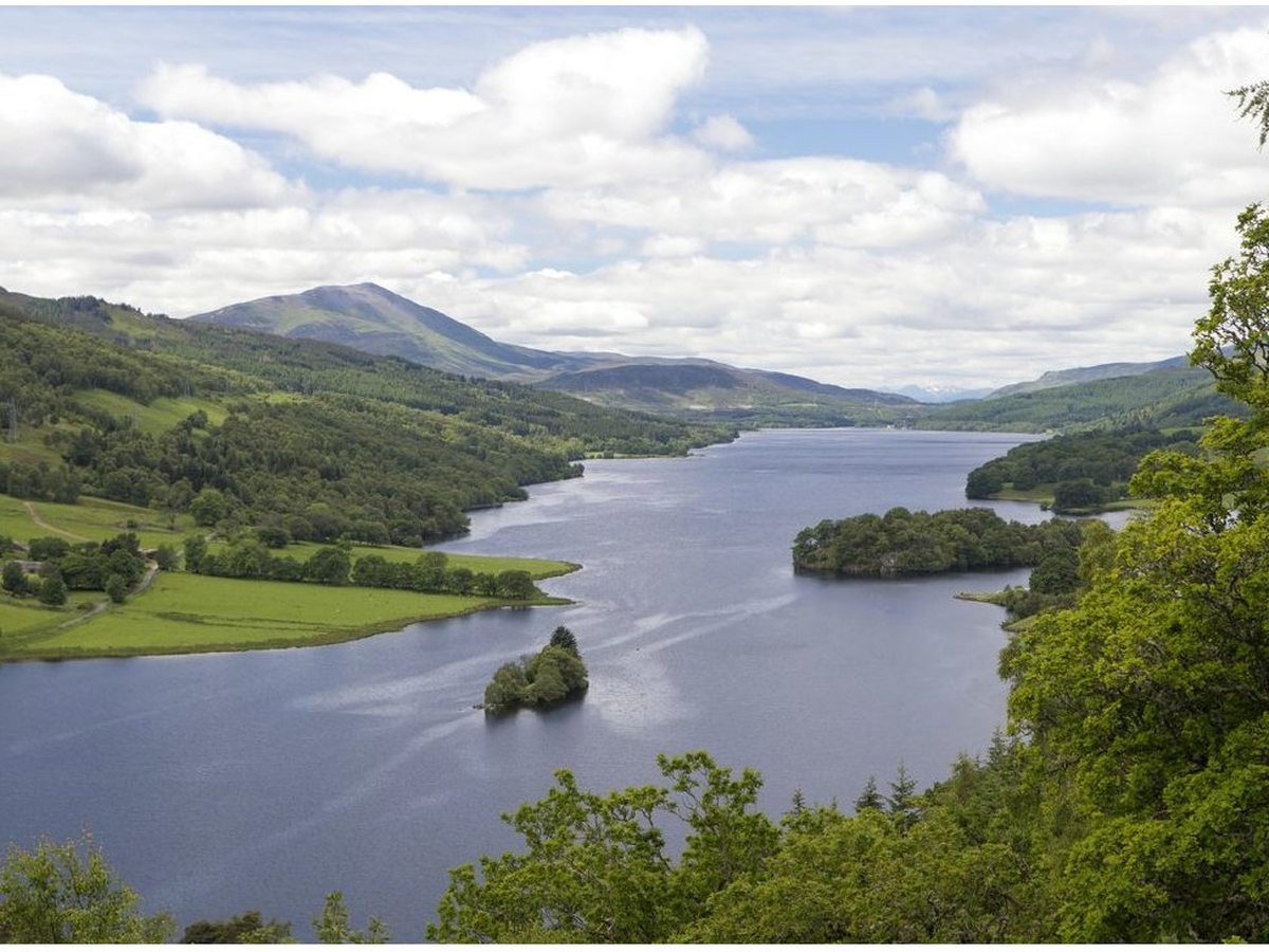 A long Highland loch seen from a high viewpoint, wooded hills on both sides and a small island near the shore, under a partly cloudy sky.