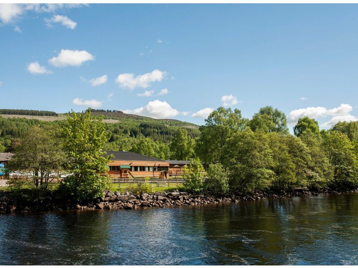 The park's timber-clad main building and lawns on the far bank of the broad River Tummel, with wooded hills behind under a blue sky.