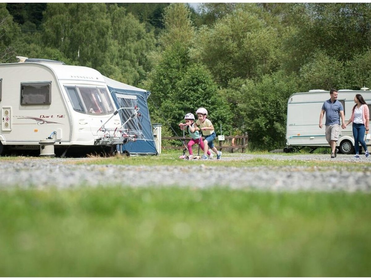 Touring caravans on grass and gravel pitches with woodland behind, a family walking between them.