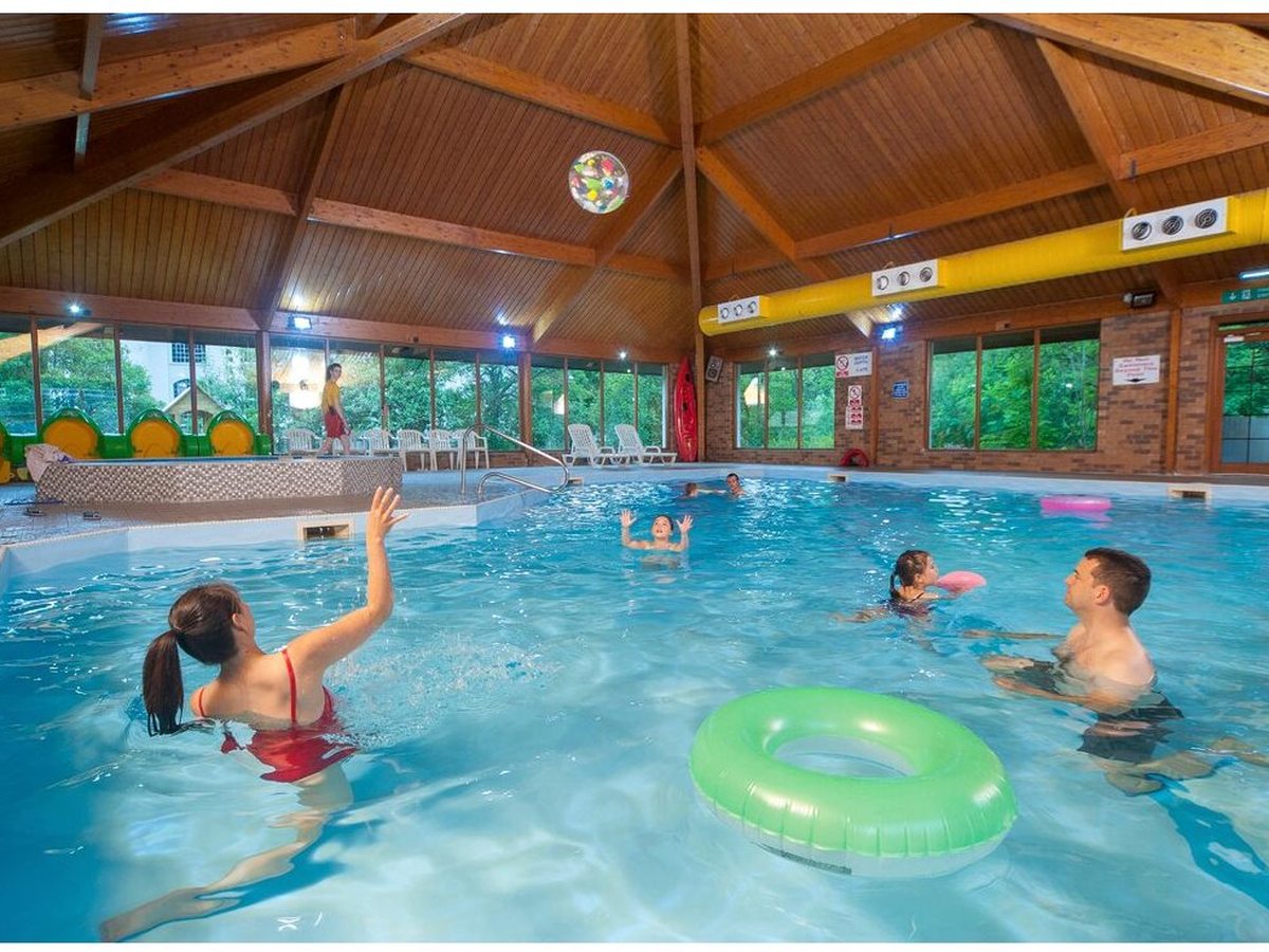 Children and adults swimming in the indoor pool, throwing a beach ball and floating a green inflatable ring, under the timber roof.