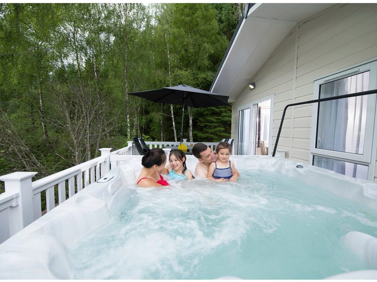 A bubbling outdoor hot tub on a lodge's decked terrace, with a woman and two children sitting in the water and woodland behind.