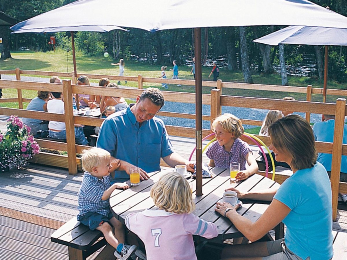 A family with young children eating at a wooden picnic table on a decked terrace under large white parasols, trees behind.